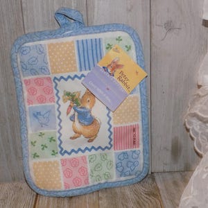 May include: A quilted pot holder featuring a patchwork design in pastel colors. The center square showcases Peter Rabbit holding carrots. The pot holder has a blue border and a hanging loop. A tag with the Peter Rabbit logo is attached.