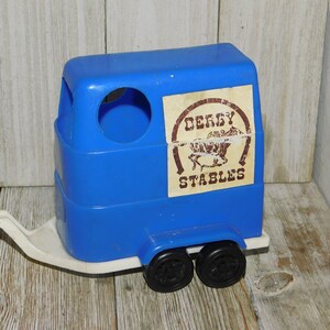 May include: Blue plastic toy horse trailer with a white hitch and black wheels. The side of the trailer has a brown label with the text "Derby Stables" and a picture of a horse.