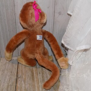 May include: A brown plush monkey toy with a pink bow on its head. The monkey has long arms and a long tail. It has a small tag on its chest. The toy is sitting on a wooden surface.