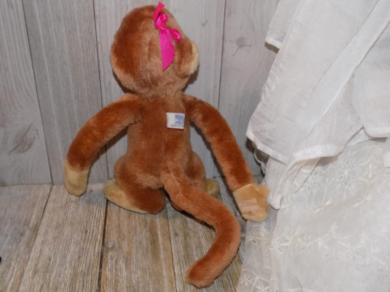May include: A brown plush monkey toy with a pink bow on its head. The monkey has long arms and a long tail. It has a small tag on its chest. The toy is sitting on a wooden surface.