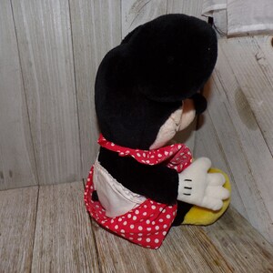 K&ouml;nnte beinhalten: Eine Pl&uuml;schpuppe von Minnie Mouse, die ein rot-wei&szlig; gepunktetes Kleid und eine wei&szlig;e Sch&uuml;rze tr&auml;gt. Die Puppe sitzt mit dem Kopf zur Seite geneigt.