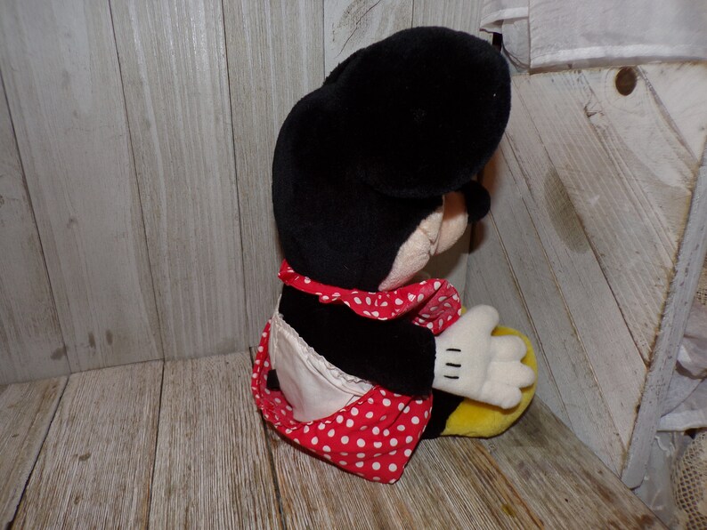 K&ouml;nnte beinhalten: Eine Pl&uuml;schpuppe von Minnie Mouse, die ein rot-wei&szlig; gepunktetes Kleid und eine wei&szlig;e Sch&uuml;rze tr&auml;gt. Die Puppe sitzt mit dem Kopf zur Seite geneigt.