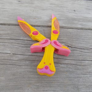May include: A pair of yellow plastic clothespins with pink accents. The clothespins are shaped like a cartoon animal with a pink nose and ears.