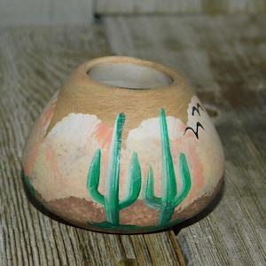 May include: A small, round, terracotta pot with a painted design of green cacti and pink clouds. The pot has a white interior and is perfect for displaying small plants or flowers.