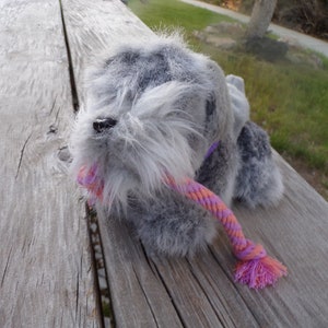 May include: A grey plush toy dog with a pink and orange scarf. The dog is lying on a wooden surface.