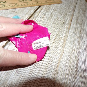 May include: A pink and blue patterned doll dress with a white tag that reads "Genuine Barbie Fashion".