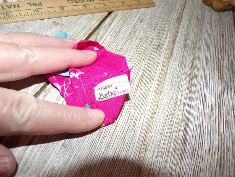 May include: A pink and blue patterned doll dress with a white tag that reads "Genuine Barbie Fashion".