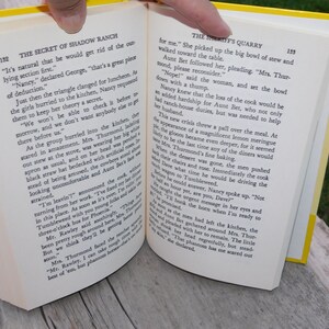 May include: Open book with yellow pages showing a scene from a novel. The text on the page reads "The Secret of Shadow Ranch" and "The Sheriff's Quarry".