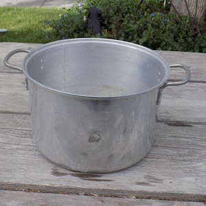 May include: A large, aluminum cooking pot with two curved handles. The pot is cylindrical with a wide opening and shows signs of wear. It sits on a weathered wooden surface, suggesting outdoor use. The pot's metallic surface reflects the surrounding environment.