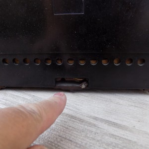 Puede incluir: Primer plano de la parte trasera de una radio vintage negra. La radio tiene una serie de peque&ntilde;as rejillas de ventilaci&oacute;n circulares a lo largo del borde inferior y una abertura rectangular. Un dedo apunta hacia la abertura.