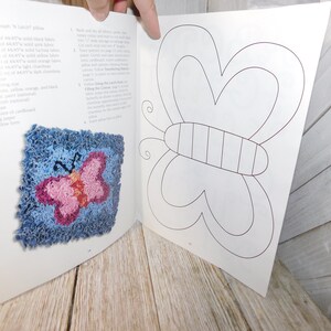 May include: A book with instructions on how to make a rug with a butterfly design. The book shows a finished rug with a blue background and a pink butterfly. The next page shows a line drawing of a butterfly.