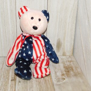 May include: A plush teddy bear with a red, white, and blue American flag pattern. The bear is sitting on a wooden surface.