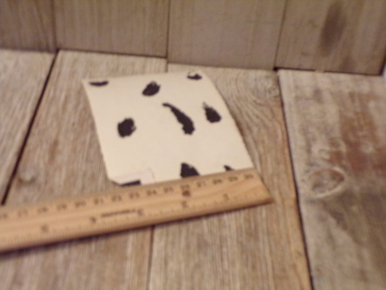May include: An off-white card with black, irregular spots. A wooden ruler, marked in inches, is placed near the card. The items are on a light-coloured wooden surface. The ruler provides a size reference.
