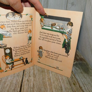 Op de afbeelding: Een open vintage kinderboek met illustraties en tekst. Het boek is getiteld "Hickery, Dickery, Dock" en toont een klok, een kat en kinderen. De tekst bevat rijmpjes over schoonmaken en eten.