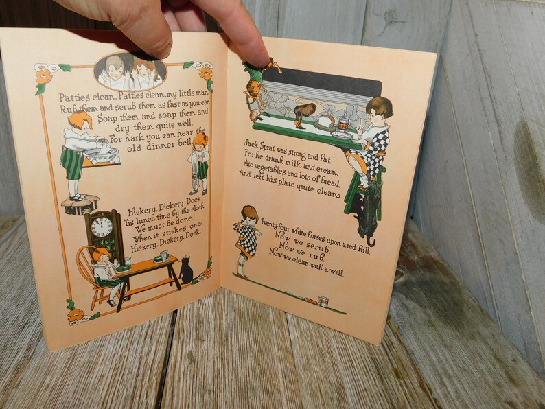 Op de afbeelding: Een open vintage kinderboek met illustraties en tekst. Het boek is getiteld "Hickery, Dickery, Dock" en toont een klok, een kat en kinderen. De tekst bevat rijmpjes over schoonmaken en eten.