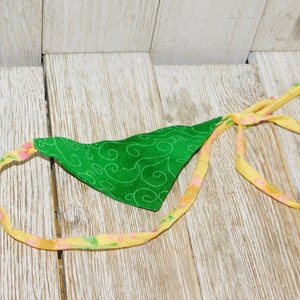 May include: A green fabric bandana with a swirl pattern and a yellow floral trim. The bandana is tied in a knot and is lying on a wooden surface.