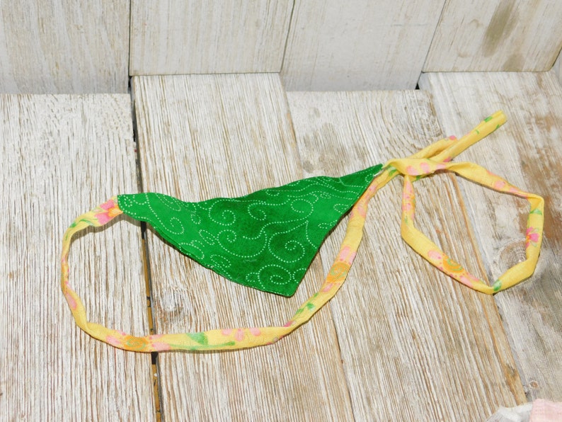 May include: A green fabric bandana with a swirl pattern and a yellow floral trim. The bandana is tied in a knot and is lying on a wooden surface.