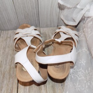 K&ouml;nnte beinhalten: Ein Paar wei&szlig;e Ledersandalen mit Schnallenverschluss und einer Korkplattformsohle. Die Sandalen haben ein Kreuzriemen-Design und sind aus einem weichen, bequemen Material gefertigt.