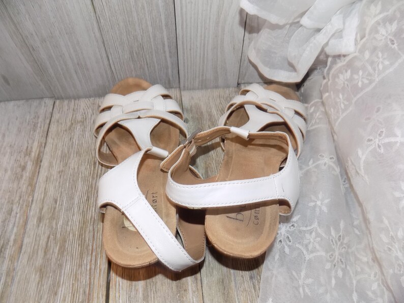 K&ouml;nnte beinhalten: Ein Paar wei&szlig;e Ledersandalen mit Schnallenverschluss und einer Korkplattformsohle. Die Sandalen haben ein Kreuzriemen-Design und sind aus einem weichen, bequemen Material gefertigt.