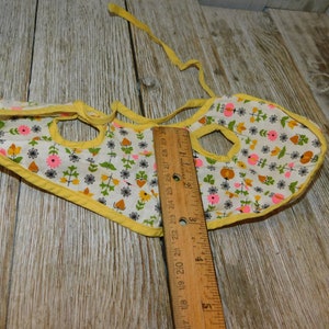 May include: A yellow bib with a white background and a floral pattern in pink, orange, and green. The bib has two holes for the neck and a tie at the back.