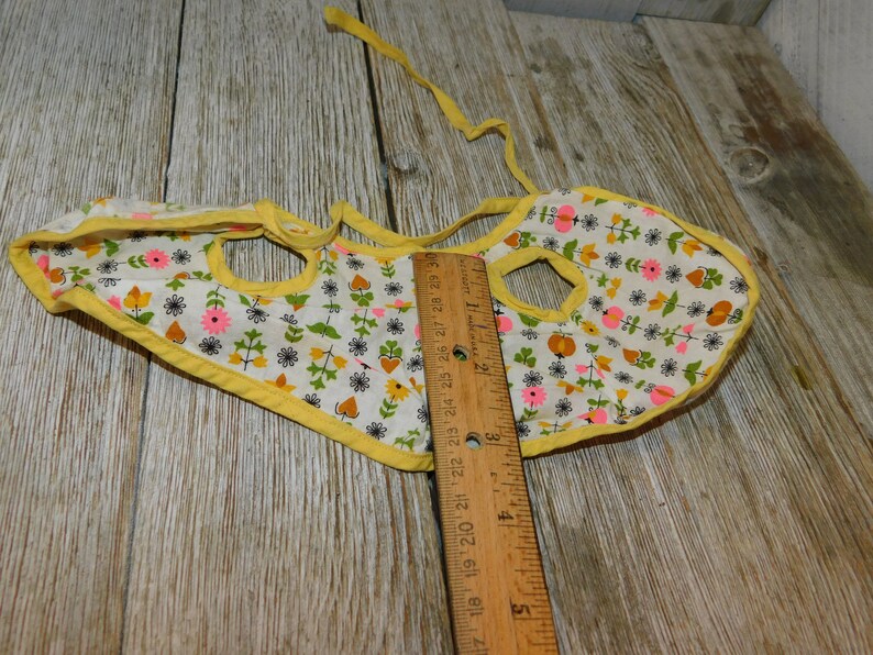May include: A yellow bib with a white background and a floral pattern in pink, orange, and green. The bib has two holes for the neck and a tie at the back.