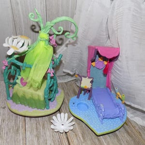 May include: Two colorful miniature playsets. One features a green and yellow structure with a white flower and pink accents. The other has a purple bed, a blue base, and a pink canopy. A white flower is also present.