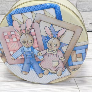 May include: A round, vintage tin with a painted illustration of two bunny dolls holding hands. The bunnies are wearing overalls and a dress, with a quilt and star background. The tin has a yellow background and a blue and white border.