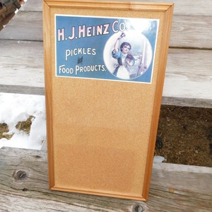 May include: A rectangular cork bulletin board framed in light wood. The top section features a vintage-style H.J. Heinz Co. advertisement with the text "H.J. Heinz Co. Pickles and Food Products" and an illustration of a person holding a jar.