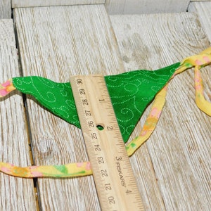 May include: A green fabric triangle with a white swirl pattern and yellow and pink trim. The triangle is attached to a yellow and pink ribbon with a ruler measuring 29 inches.