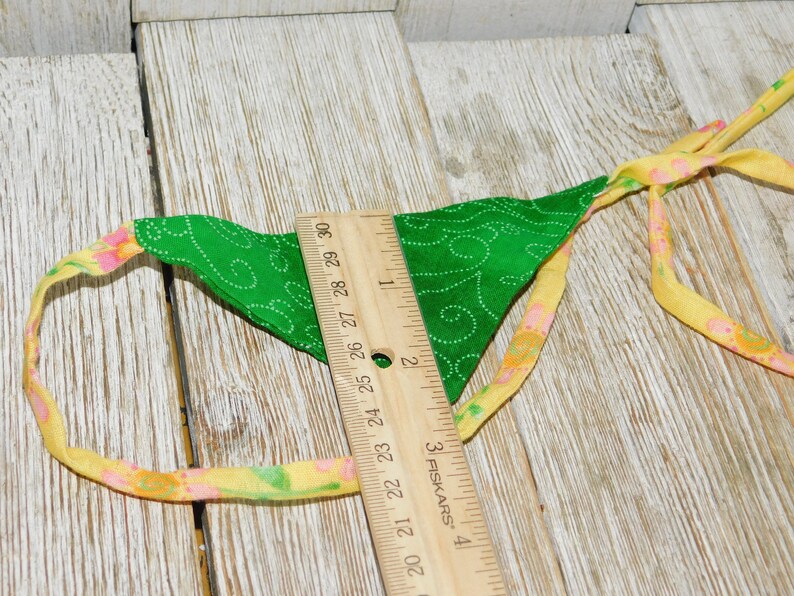 May include: A green fabric triangle with a white swirl pattern and yellow and pink trim. The triangle is attached to a yellow and pink ribbon with a ruler measuring 29 inches.