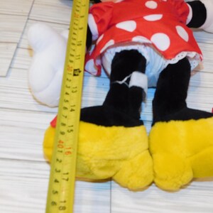 Op de afbeelding: Een pluche Minnie Mouse pop met een rode jurk met witte stippen, zwarte benen en gele schoenen. De pop is ongeveer 40 cm hoog, gemeten met een gele meetlint.