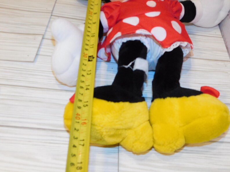 Op de afbeelding: Een pluche Minnie Mouse pop met een rode jurk met witte stippen, zwarte benen en gele schoenen. De pop is ongeveer 40 cm hoog, gemeten met een gele meetlint.