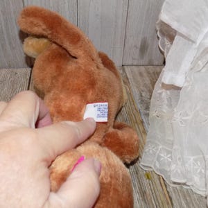 May include: A brown plush bunny toy with long ears. The toy has a tag attached. A hand is touching the tag. A white lace fabric is in the background.