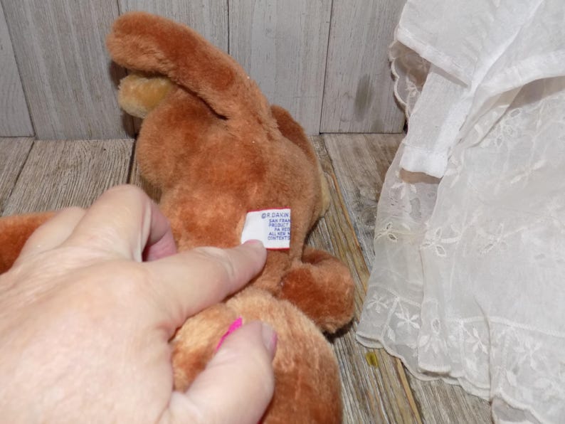 May include: A brown plush bunny toy with long ears. The toy has a tag attached. A hand is touching the tag. A white lace fabric is in the background.