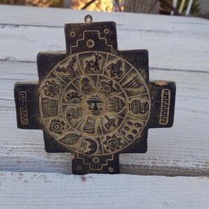 May include: A dark brown, cross-shaped wall plaque featuring a central circular relief carving. The design includes a human face at the center surrounded by various symbols, including animals, plants, and geometric patterns, with a small metal hanging loop.