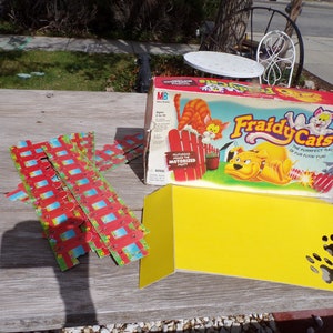 May include: A vintage board game called "Fraidy Cats" with a yellow box and a red, white, and blue game board. The game features a motorized cat and a dog chasing the cats. The game is in good condition and includes all of the pieces.