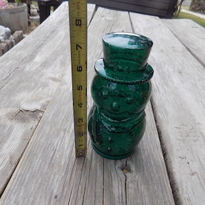 May include: A green glass snowman figurine with a sparkly finish. The snowman has a round body, a black hat, and a white face with black eyes and a red mouth.