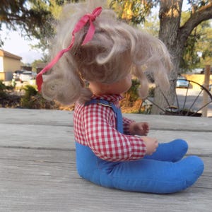 Puede incluir: Una mu&ntilde;eca vintage con cabello rubio y un lazo rosa. Lleva una camisa a cuadros rojos y blancos y un mono azul.