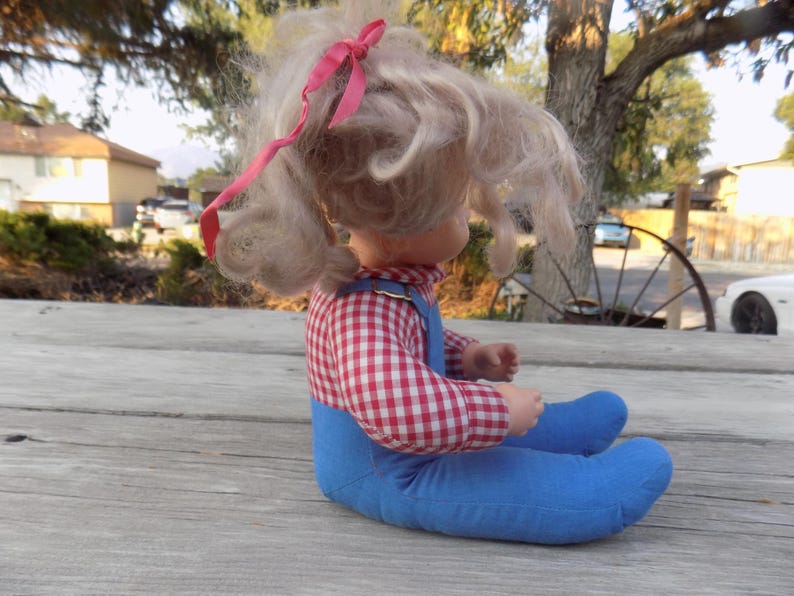 Puede incluir: Una mu&ntilde;eca vintage con cabello rubio y un lazo rosa. Lleva una camisa a cuadros rojos y blancos y un mono azul.