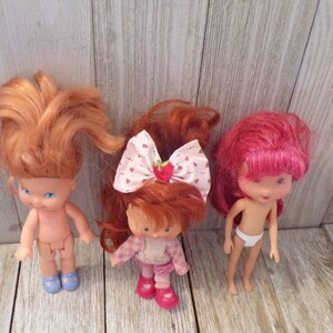 Pode incluir: Tr&ecirc;s bonecas Strawberry Shortcake com cabelo ruivo. Uma boneca est&aacute; vestindo uma camisa xadrez rosa e branca e cal&ccedil;as rosa. Outra boneca est&aacute; vestindo uma fralda branca. A terceira boneca est&aacute; nua.