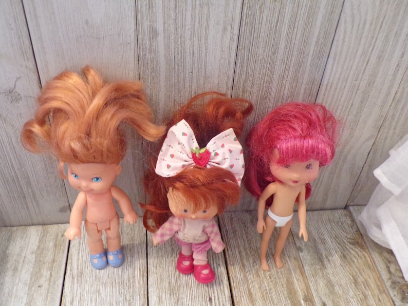 Pode incluir: Tr&ecirc;s bonecas Strawberry Shortcake com cabelo ruivo. Uma boneca est&aacute; vestindo uma camisa xadrez rosa e branca e cal&ccedil;as rosa. Outra boneca est&aacute; vestindo uma fralda branca. A terceira boneca est&aacute; nua.