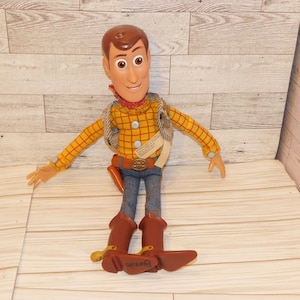 May include: A Woody doll from the Toy Story franchise. The doll has a brown head of hair, a red bandana, a yellow plaid shirt, blue jeans, and brown boots. The doll is wearing a vest and a belt with a buckle.