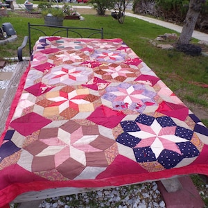 May include: A large, hand-stitched quilt with a starburst pattern. The quilt is made of various shades of pink, red, brown, and white fabrics. It is laid out on a wooden table with a gravel surface.