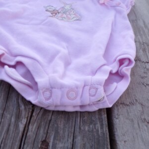 May include: A light purple baby onesie with a white bunny and flower embroidery. The onesie has three white snaps at the bottom.