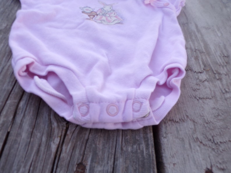 May include: A light purple baby onesie with a white bunny and flower embroidery. The onesie has three white snaps at the bottom.
