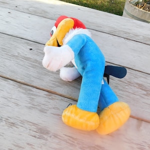 May include: A blue and yellow plush toy of a cartoon woodpecker with a red head and white feathers. The toy is lying on a wooden surface.