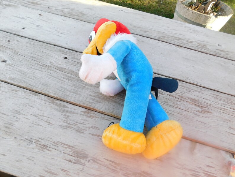 May include: A blue and yellow plush toy of a cartoon woodpecker with a red head and white feathers. The toy is lying on a wooden surface.