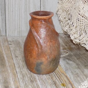 May include: A small, brown, hand-crafted ceramic vase with a unique, twisted design. The vase has a rustic, earthy appearance.