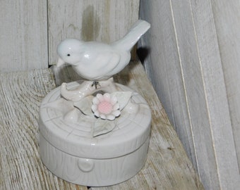 May include: A white porcelain bird figurine perched on a branch with a pink flower. The bird is sitting on a round, white porcelain box with a woodgrain design.