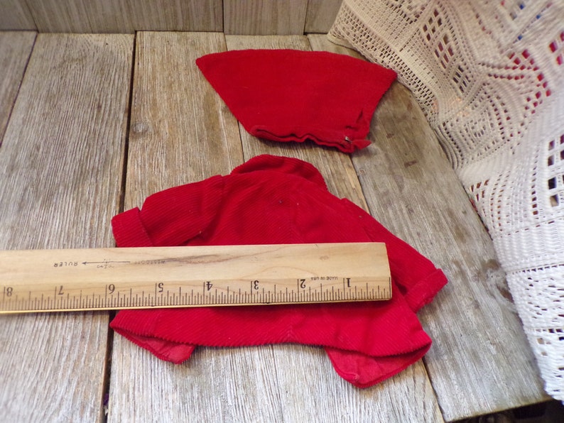 K&ouml;nnte beinhalten: Zwei rote Cordjacken f&uuml;r eine Puppe. Die gr&ouml;&szlig;ere Jacke liegt flach ausgelegt, die kleinere Jacke ist auf einem Holzlineal gefaltet. Das Lineal ist in Zoll eingeteilt.